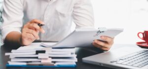 Mężczyzna przeglądający dokumenty na podkładce przy stosie papierów i laptopie na biurku – kontrola skarbowa w firmie